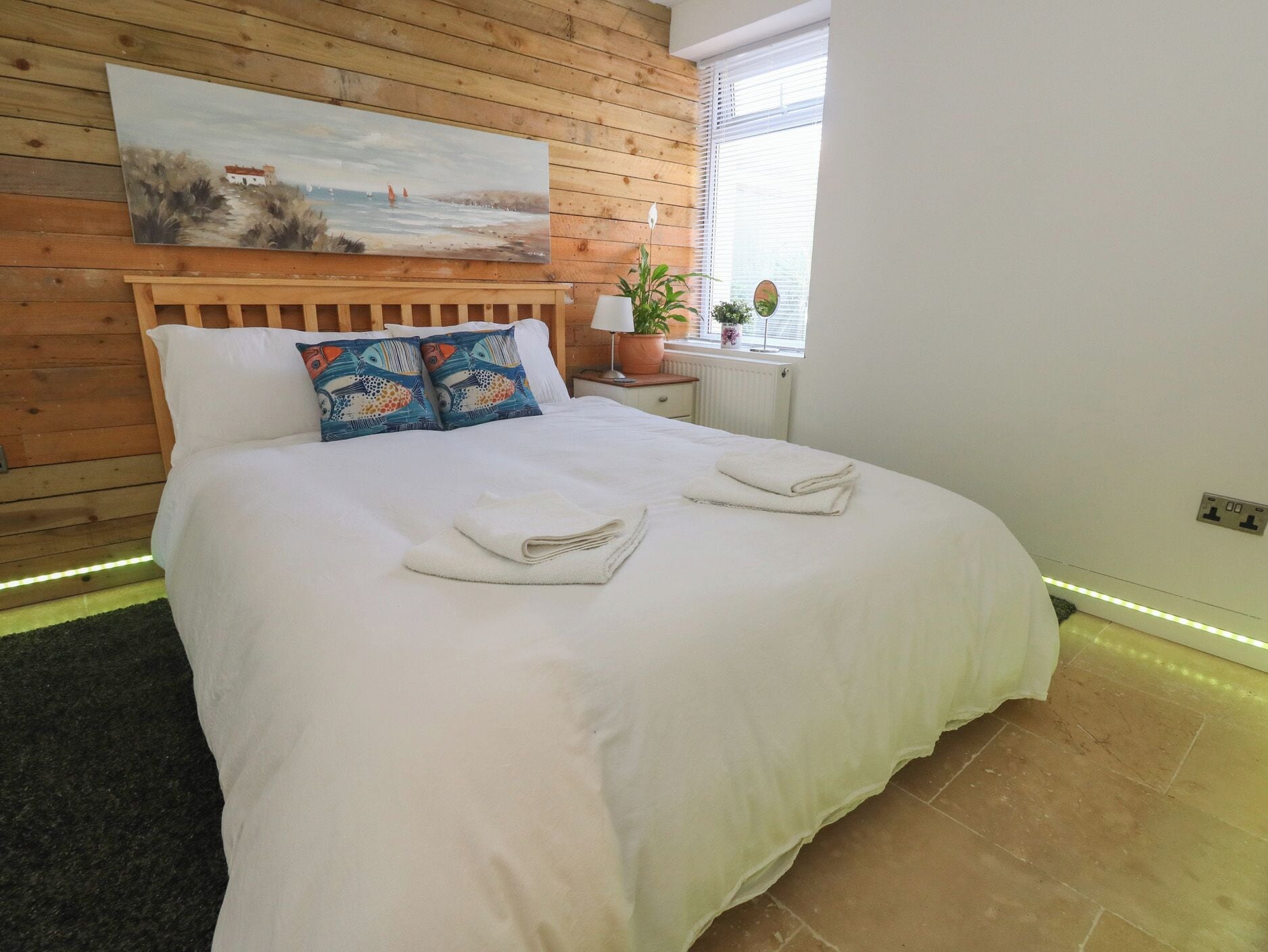 A neatly made bed with white linens and towels, a wooden headboard, a painting of a coastal scene, and a window with blinds.