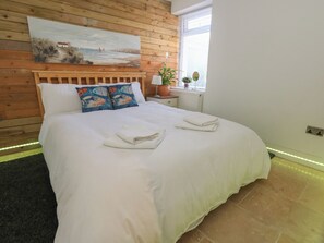 A neatly made bed with white linens and towels, a wooden headboard, a painting of a coastal scene, and a window with blinds. - Tipsy Turtle (Portland)