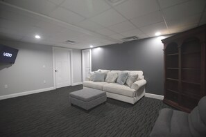 A room with a grey carpet, a white sofa with patterned cushions, a grey ottoman, a wooden cabinet, and a flat-screen TV mounted on the wall. - ELITE SUITES  5 BEDROOM RETREAT (Pittsburgh)