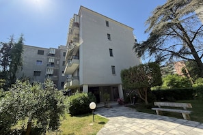 Um edifício residencial de vários andares, com varandas, um pátio pavimentado e vegetação ao redor.