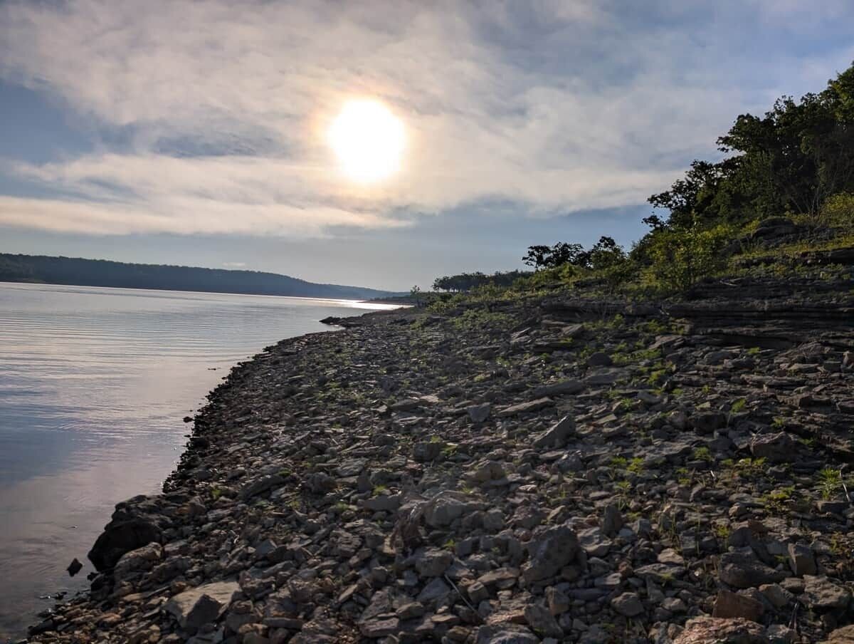 A rocky shoreline with calm water, a clear sky, and distant land.