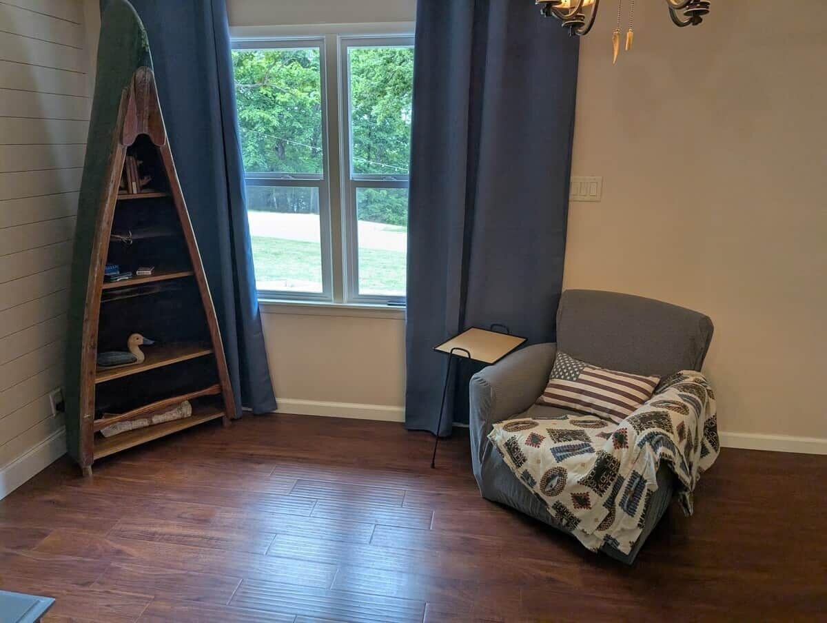 Une pièce avec une échelle en bois, un fauteuil gris, une petite table et une fenêtre avec des rideaux.