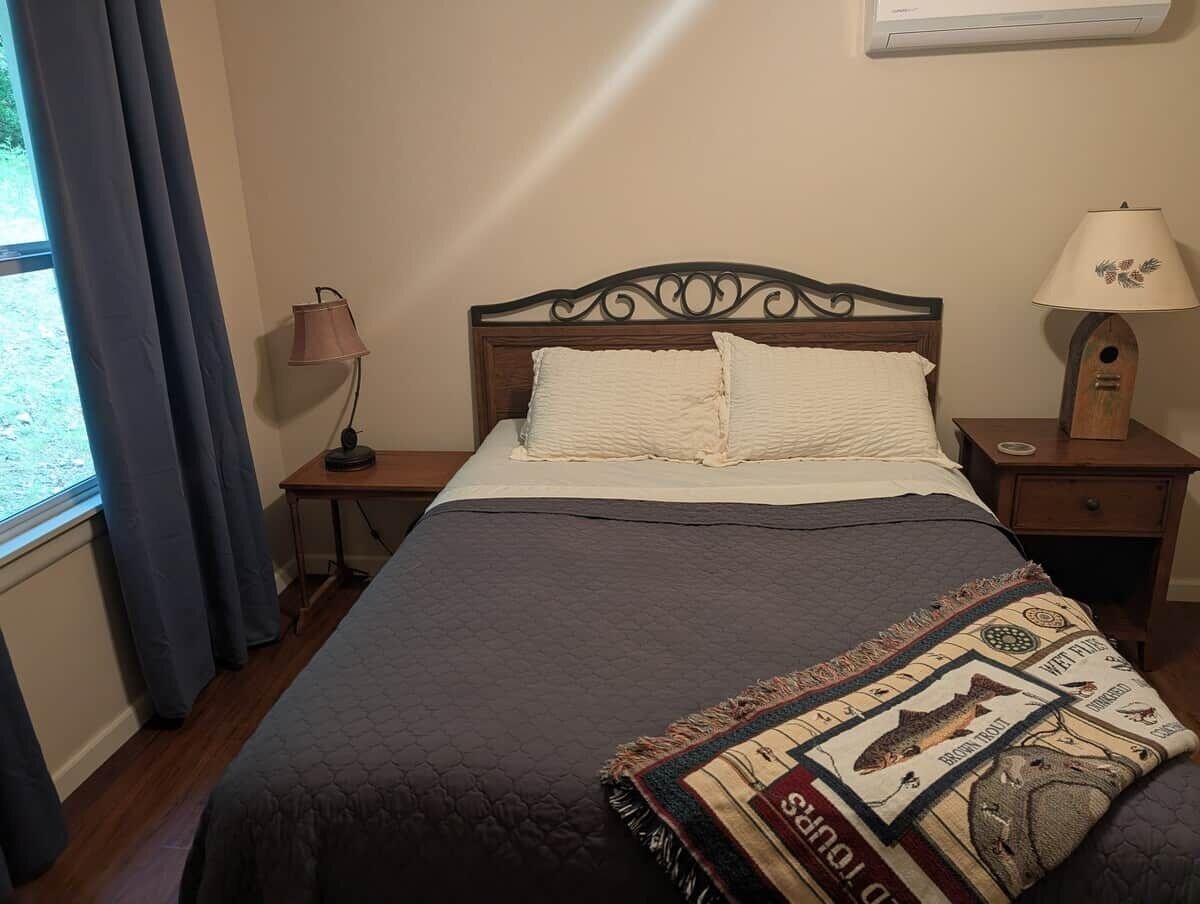 Un lit avec une couverture sombre, deux oreillers, un plaid décoratif, une lampe de chevet et une table de chevet avec une lampe.