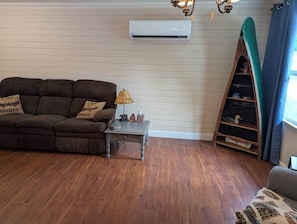 A living room with a brown sofa, a small table with a lamp, an air conditioning unit on the wall, and a wooden shelf with decorative items.