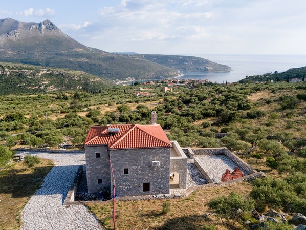 Un edificio de piedra con techo de tejas rojas, rodeado por un patio de piedra, con el paisaje montañoso y la costa como telón de fondo.