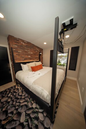 Quarto com parede de destaque em tijolo, cama com roupa de cama branca, um travesseiro laranja, cabeceira e piso de madeira.