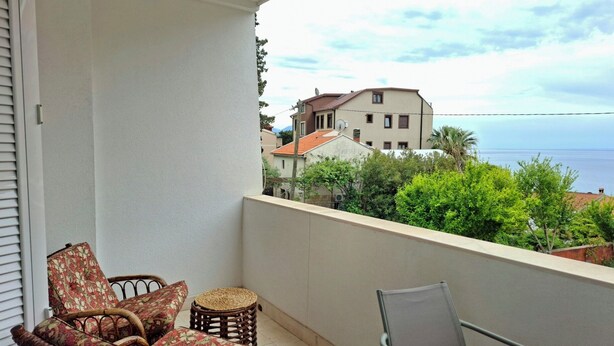 Un balcon avec des fauteuils en osier et une table, d’où l’on peut admirer un quartier résidentiel et la mer.
