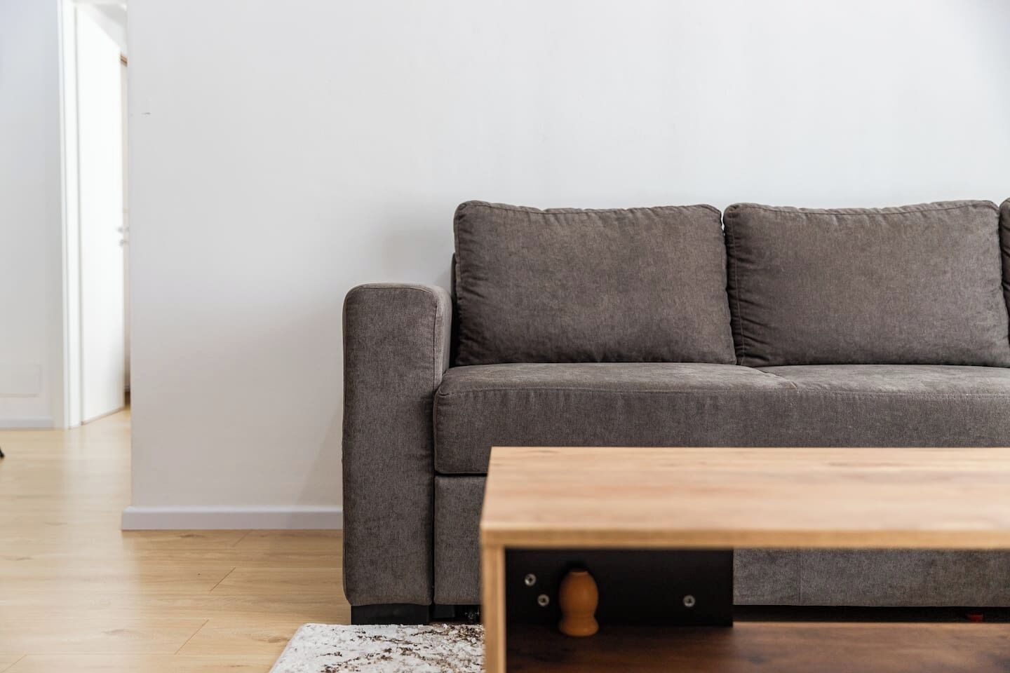 Un canapé gris, accompagné d’une table basse en bois, se trouve dans une pièce dont le sol est recouvert de parquet et les murs sont peints en blanc.