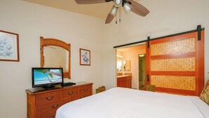 Ein Schlafzimmer mit einem Bett, einem Fernseher auf einer Holzkommode, einem Deckenventilator, einem Spiegel und einer Schiebetür, die zu einem Badezimmer führt.