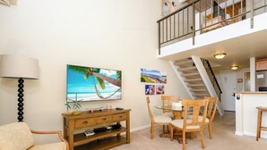 Ein Wohnzimmer mit Fernseher, Holzmöbeln, einem Esstisch und einer Treppe, die zu einer oberen Ebene führt.