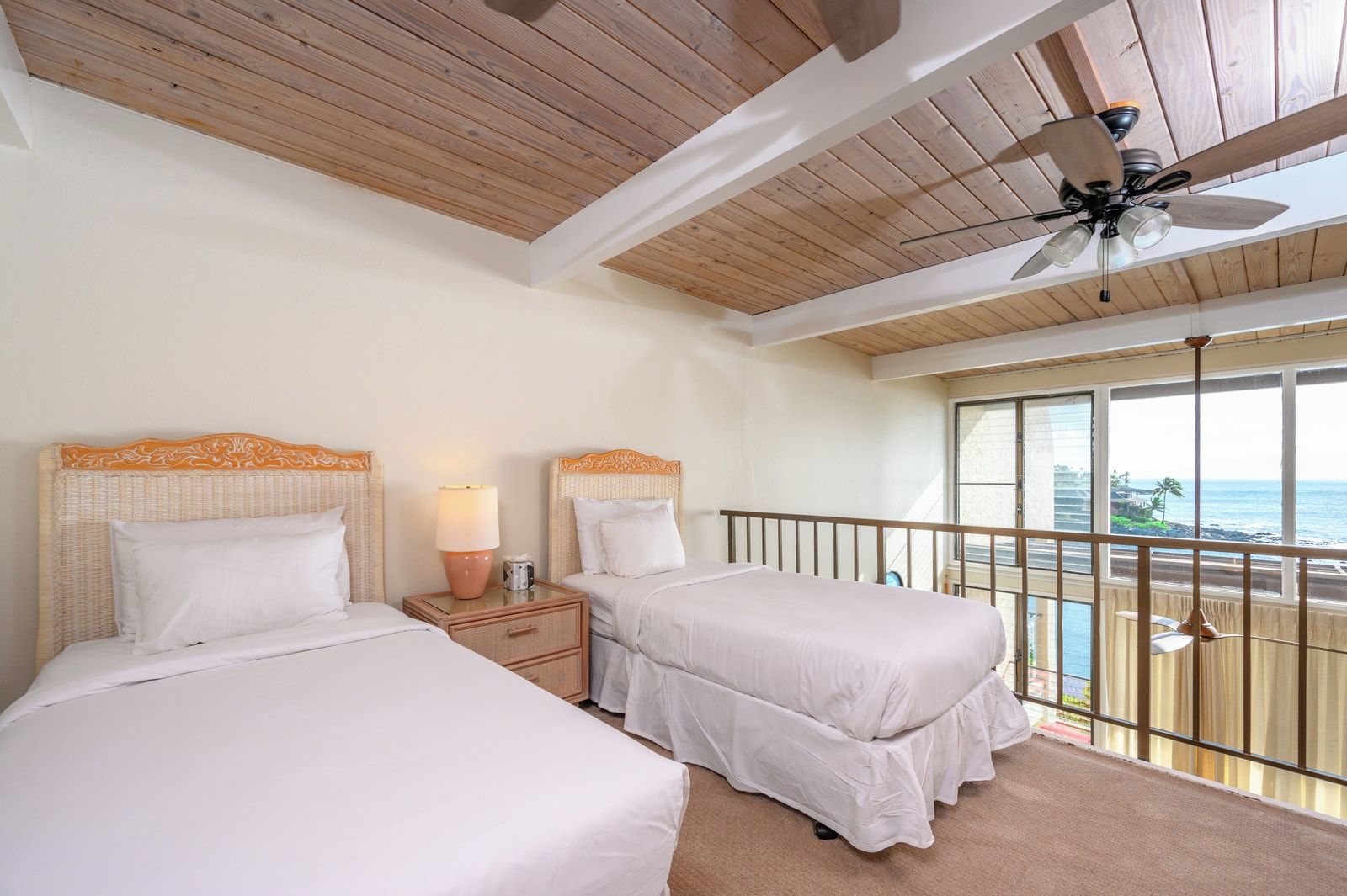 Habitación de hotel con dos camas, ventilador de techo, mesita de noche con lámpara y balcón con vista al océano.