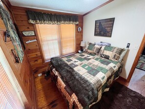 A wooden cabin bedroom with a bed, a nightstand, a lamp, a window with blinds, and a wall clock.