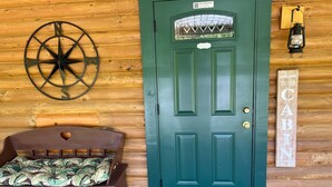 Interior de una cabaña de madera con una puerta verde, un adorno de pared con brújula, una cama con un colchón estampado con hojas y un letrero de bienvenida.