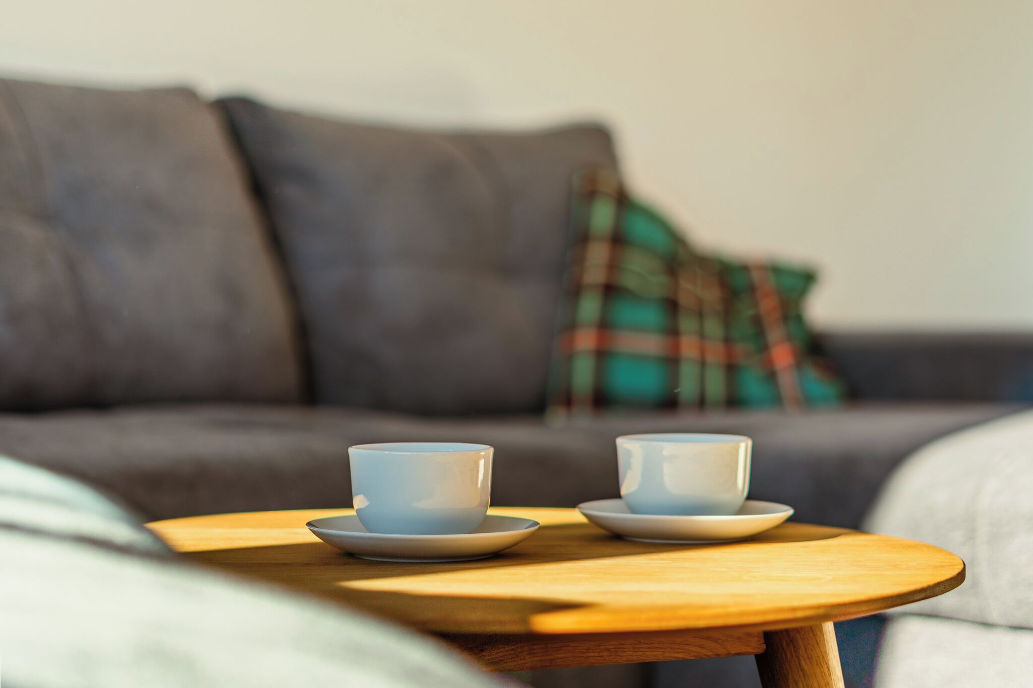 Una mesa de madera con dos tazas y platillos blancos, un sofá gris con un cojín escocés, y un cojín blanco en una sala de estar.