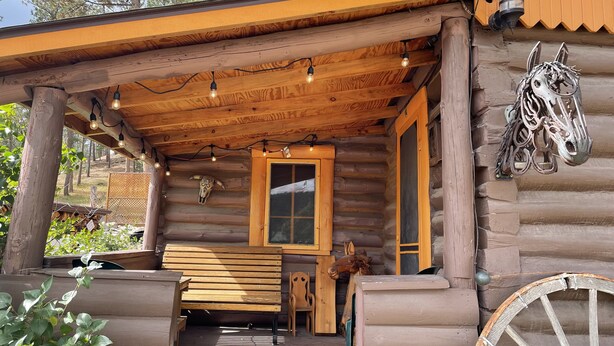 Una cabaña de troncos con un porche de madera, luces de cuerda, una decoración con forma de cabeza de caballo y un banco de madera.