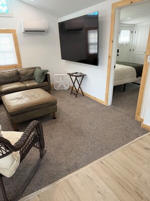 Ein Wohnzimmer mit einer Couch, einem kleinen Tisch, einem an der Wand montierten Fernseher und einem Bett im Hintergrund.