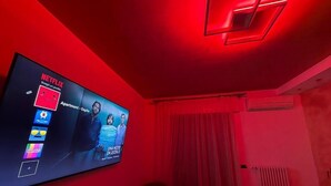 Ett rum med en tv-skärm som visar Netflix-gränssnittet, ett rött stämningsljus, ett element och ett fönster med gardiner.