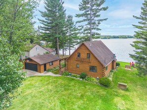 A log cabin with a deck and a garage, surrounded by a grassy yard and trees, overlooking a lake.