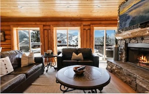 Ein Blockhaus-Interieur mit Kamin, Ledersofas, einem Couchtisch und Blick auf verschneite Berge.