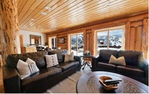 Ein Interieur einer Blockhütte mit zwei Ledersofas, einem Couchtisch und Blick auf eine verschneite Landschaft durch große Fenster.