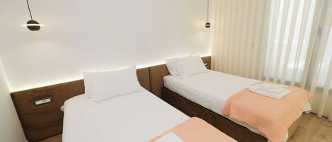 A hotel room with two beds, a wooden headboard, a bedside table, and a window with sheer curtains.