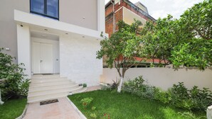 Un bâtiment moderne à l’extérieur blanc, un escalier menant à une porte, un petit jardin avec de la pelouse et des fleurs, et un arbre.