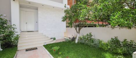 A modern building with a white exterior, a set of stairs leading to a door, a small garden with grass and flowers, and a tree.