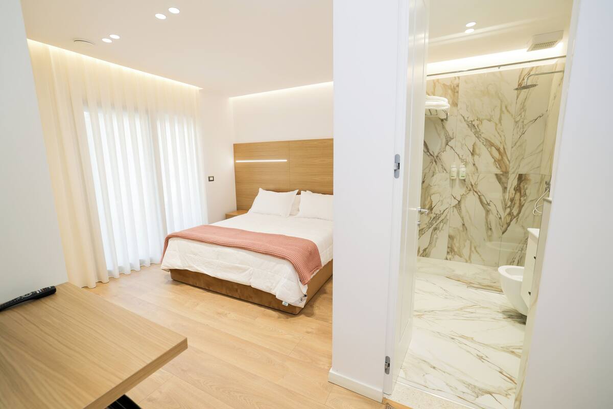Habitación de hotel moderna con cama de madera, una mesita de noche, baño con ducha de mármol y un amplio ventanal con cortinas.