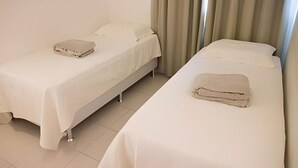 Due letti singoli con lenzuola bianche e un asciugamano piegato su ciascuno, una parete spoglia e una tenda sul lato destro.