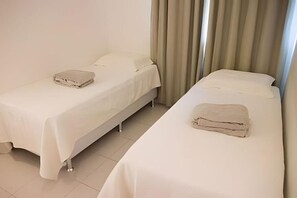 Zwei Einzelbetten mit weißen Laken und einem zusammengeklappten Handtuch, eine schlichte Wand und ein Vorhang auf der rechten Seite.
