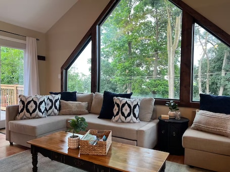 A living room with a sofa, coffee table, and large windows offering a view of trees.. Blue Lakeview Home