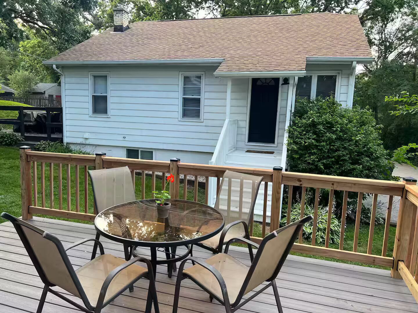 A deck with a glass-top table and four chairs, a wooden railing, and a house with a brown roof and white siding in the background.