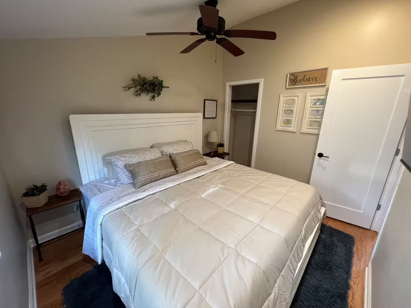 Une chambre à coucher avec un lit, un ventilateur de plafond, une porte et une petite plante posée sur une table de chevet.