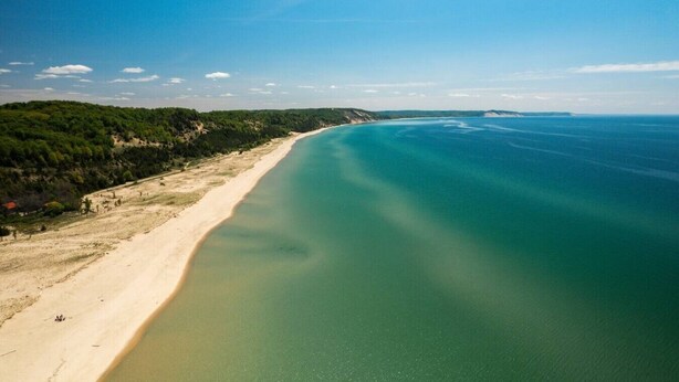 Et kystlandskab med en sandstrand, et klart blå hav og en skovklædt bakke.