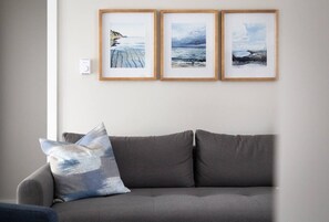 A grey sofa with a single pillow, three framed artworks on the wall, and a thermostat.