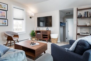 A modern living room with a flat-screen TV, a wooden coffee table, a grey sofa, a wicker chair, and a bookshelf.