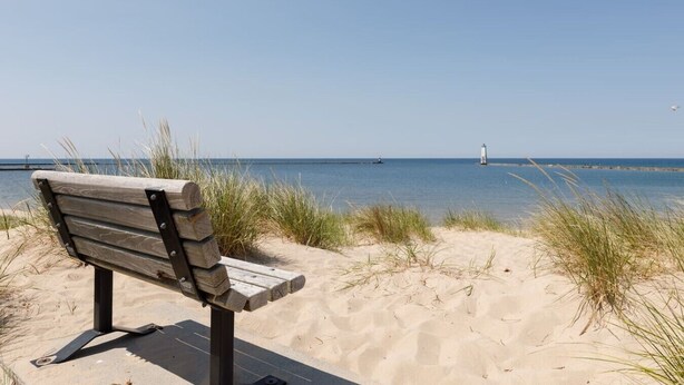 Eine Bank an einem sandigen Strand mit freiem Blick auf das Meer und einem Leuchtturm in der Ferne.