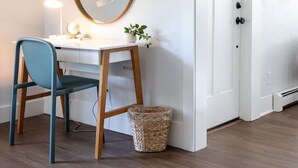 Ein Zimmer mit Schreibtisch, Stuhl, Spiegel, Lampe, Zimmerpflanze und einer Tür.