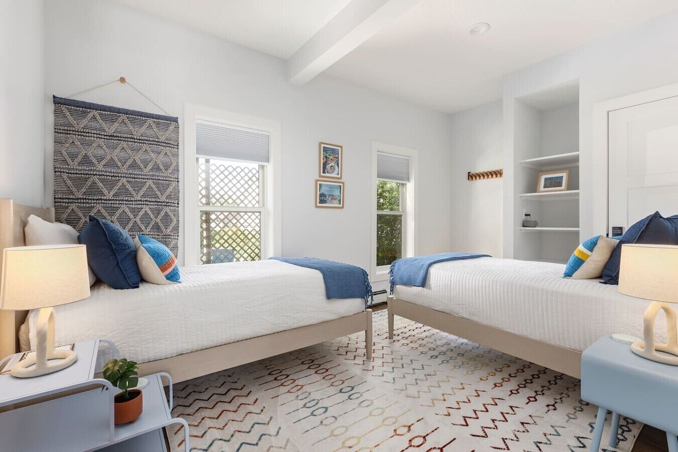A bedroom with two beds, a desk, a chair, a lamp, a plant, and a patterned rug.