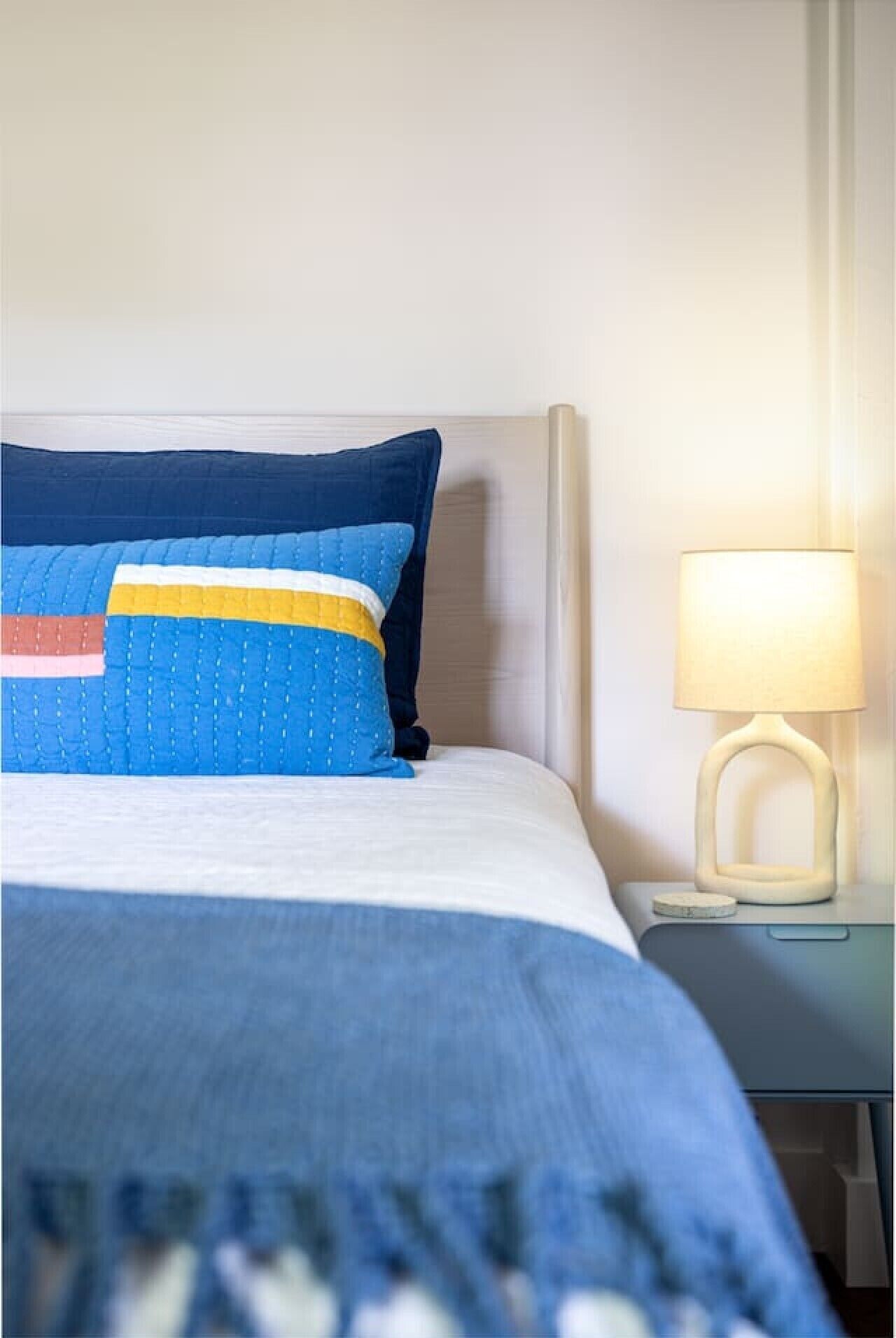 Ein ordentlich bezogenes Bett mit blauer und weißer Bettdecke und Kissen, ein Nachttisch mit einer Lampe sowie eine schlichte Wand.
