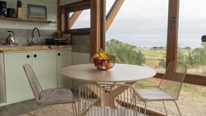 A kitchen with a round table and chairs, a fruit bowl on the table, and a view of the countryside through large windows. - La Vertiente Pullay (Cobquecura)