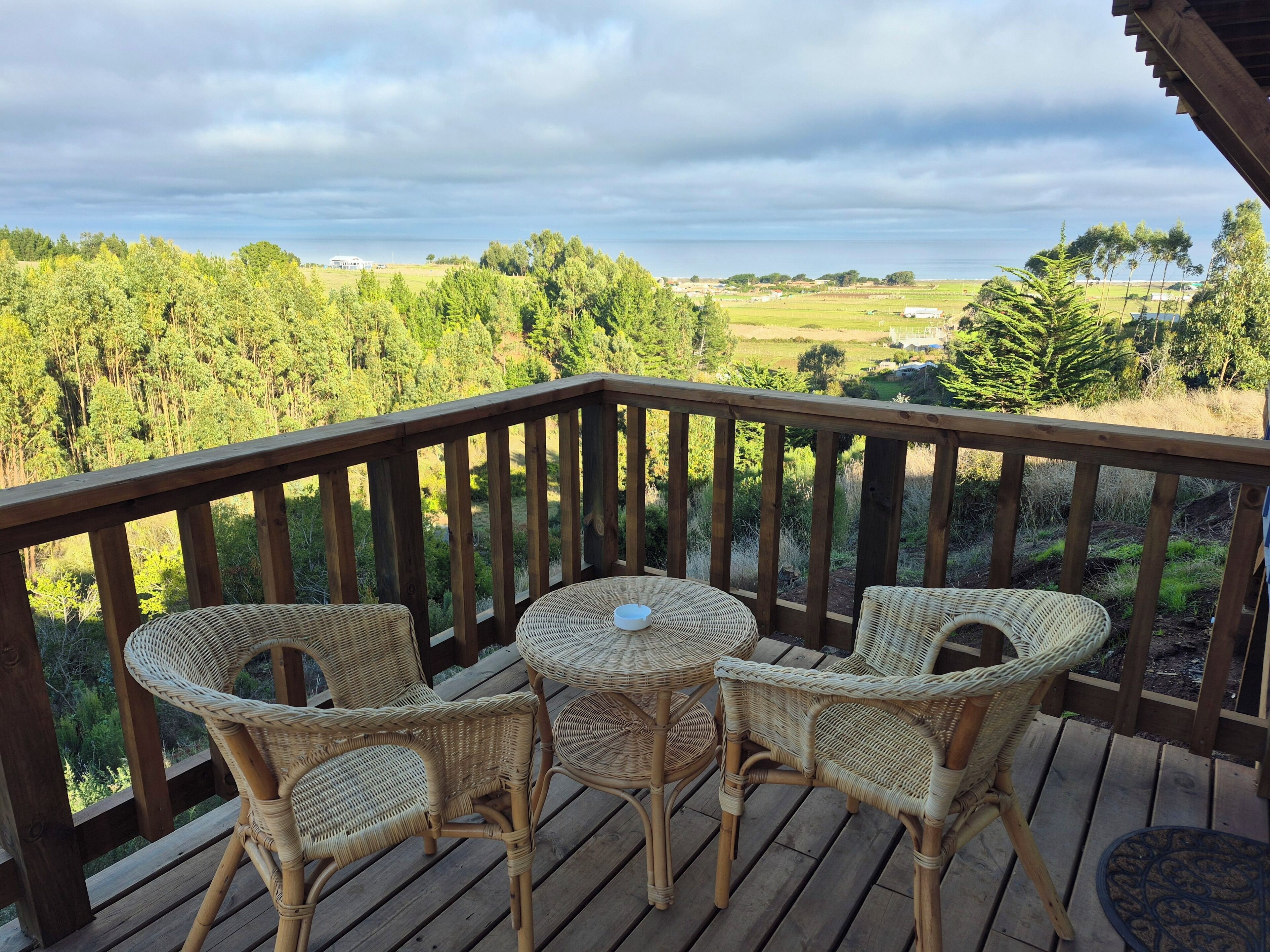 A wooden deck with wicker chairs and a small round table, overlooking a green landscape with trees and fields.
