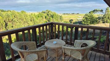 A wooden deck with wicker chairs and a small round table, overlooking a green landscape with trees and fields.