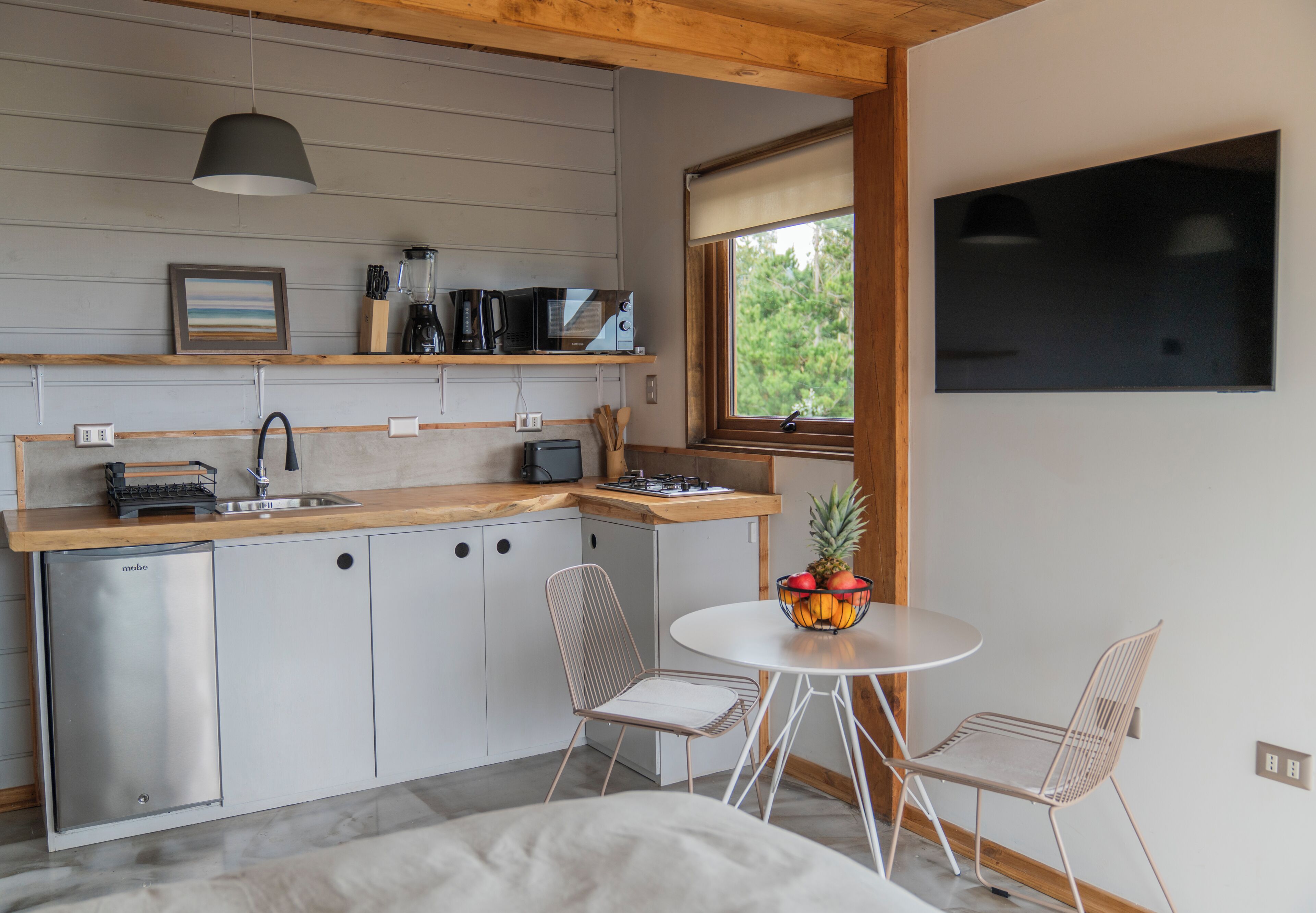 Een compacte keuken met een vaatwasser, magnetron en koffiezetapparaat, een eethoek met een tafel en twee stoelen, en een groot raam met uitzicht op groen.