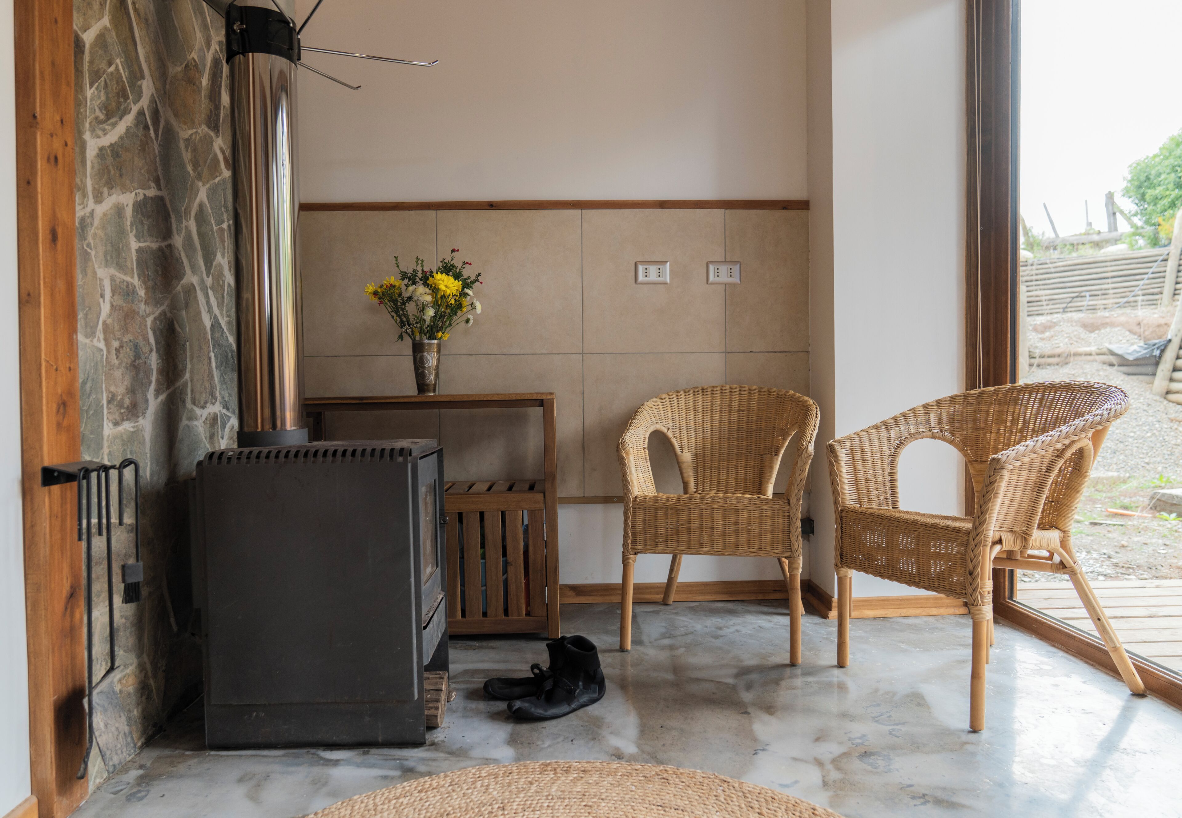 A room with a stone wall, a black heater, wicker chairs, a wooden table with a vase of flowers, and a window with a view of the outdoors.