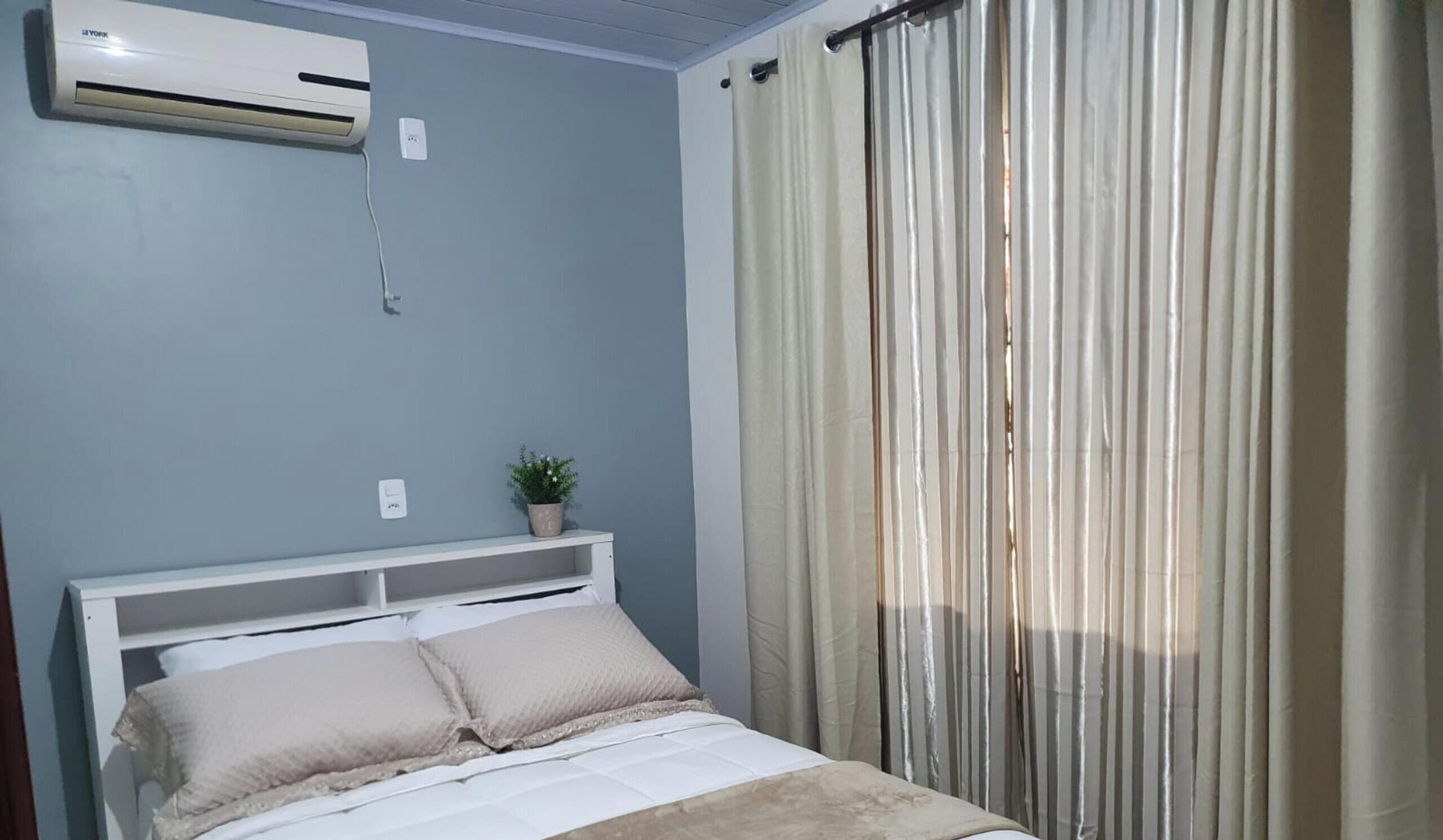 A bedroom with a bed, an air conditioner, a potted plant, and a window with curtains.