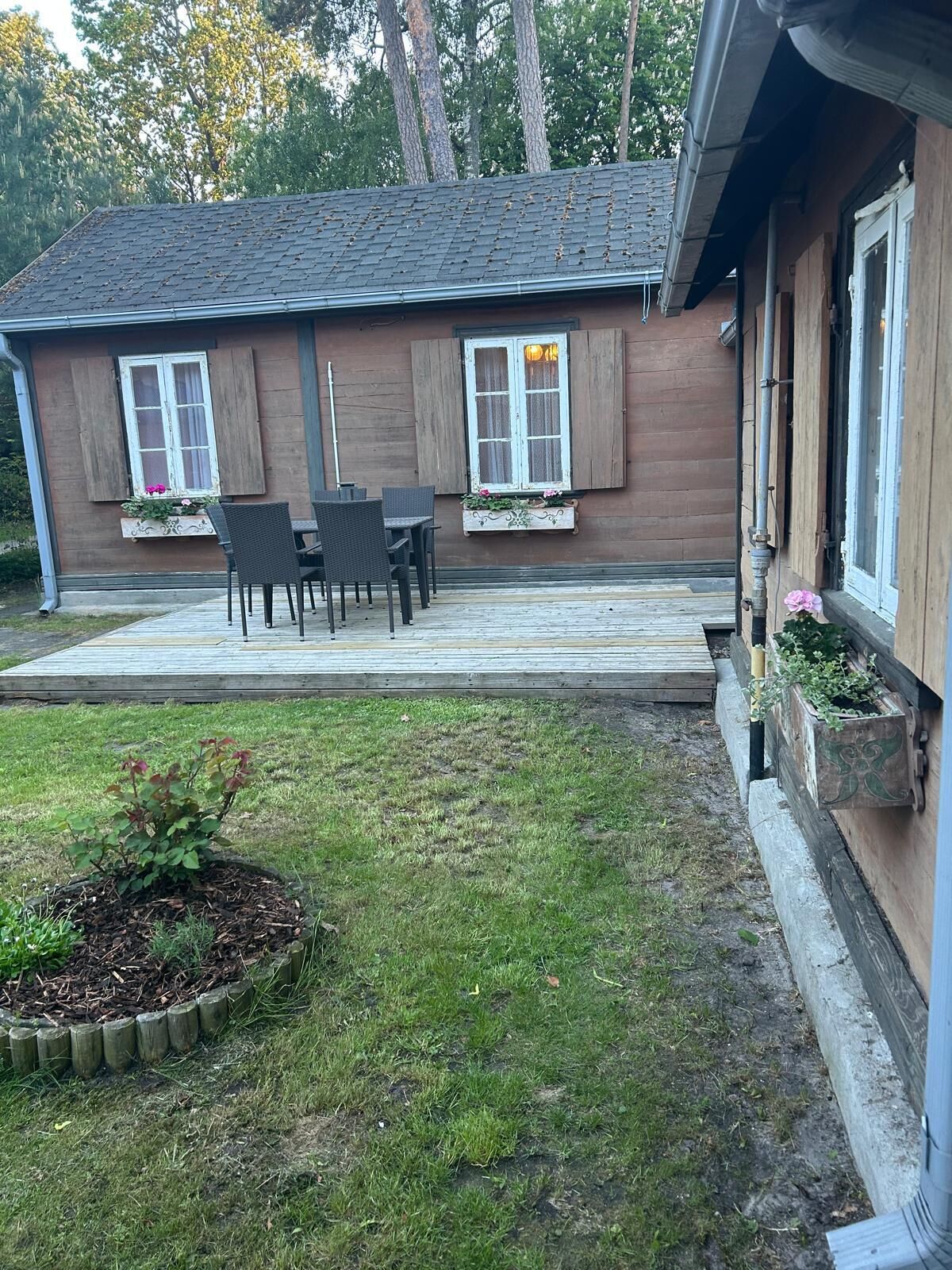 A wooden house with a patio, two windows with shutters, and a small garden area.