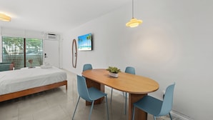 Ett modernt sovrum med en säng, ett matbord med stolar, en krukväxt och en tavla med strandmotiv på väggen.