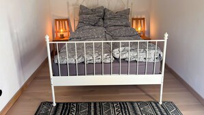 Una cama de metal blanco con una colcha y almohadas estampadas, dos lámparas de noche y una alfombra blanco y negro sobre un piso de madera.