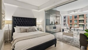 Ein modernes Schlafzimmer mit einem großen Bett, einer Glastrennwand, Blick auf eine Stadtlandschaft und einem Balkon mit Sitzgelegenheit.